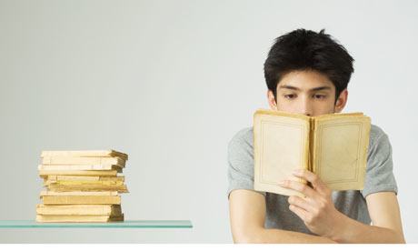 Stock image of a young man reading a book beside a pile of books