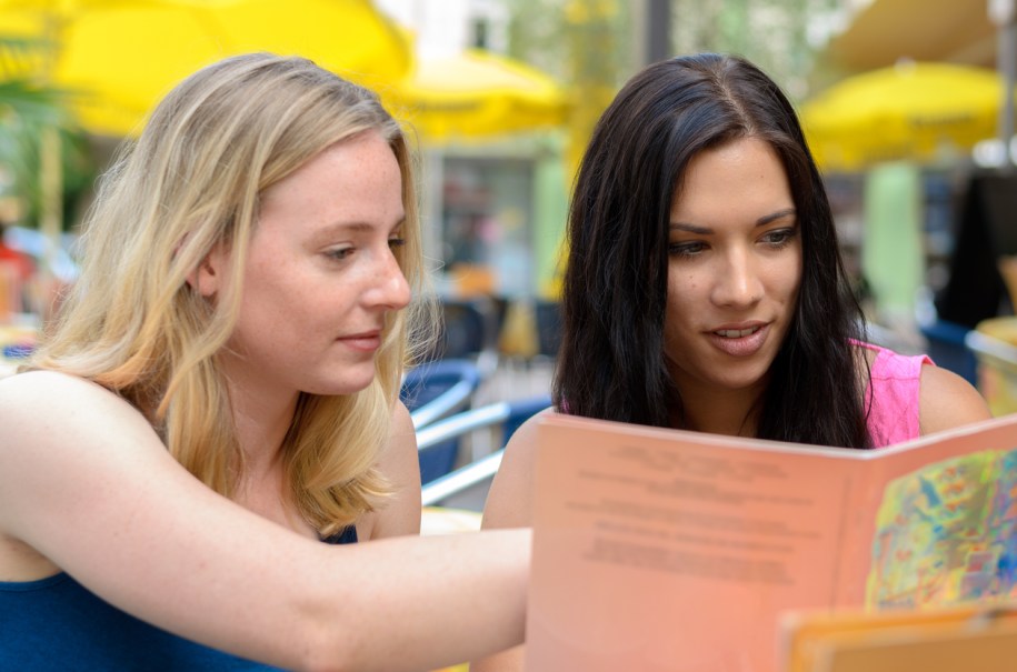 Pair of beautiful young adult women looking at book or menu together at outdoor cafe
