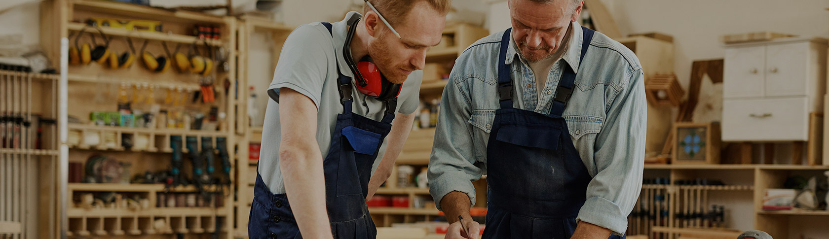 Alt text: Two men in aprons work together in a workshop, with one leaning over a workbench and the other standing beside him. Shelves of tools and supplies fill the background, suggesting a hands-on training environment.